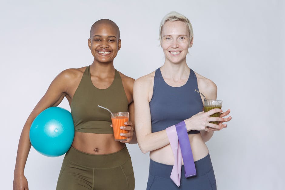 Positive multiracial sportive women in sportswear standing with fitness ball and resistance bands and drinking smoothies after workout against white background